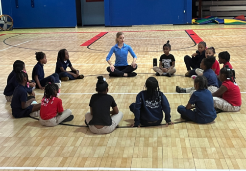 Children sit in a circle with their instructor in a school gym.