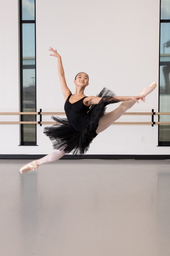 A girl in a black tutu leaps in the air in the studio.