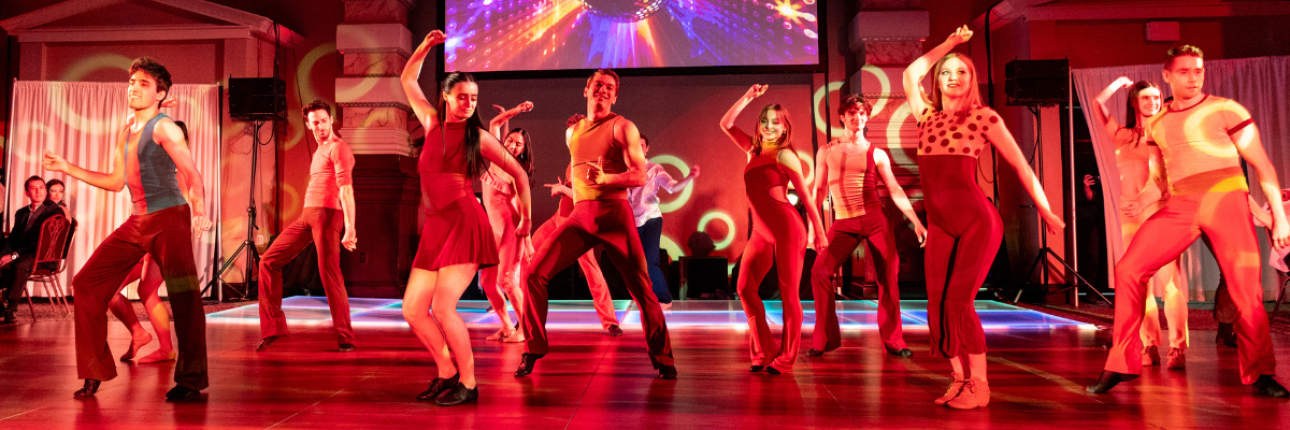 Dancers performing in blue leotards and black bottoms, with the stage lit by a red light