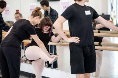 A Tour de Force student stretches at the barre alongside a dancer.